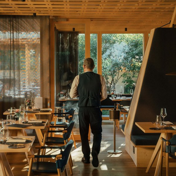 Sensoria Soulmates – Creiamo ricordi felici. Cameriere cammina in ristorante moderno con mobili in legno e grandi finestre