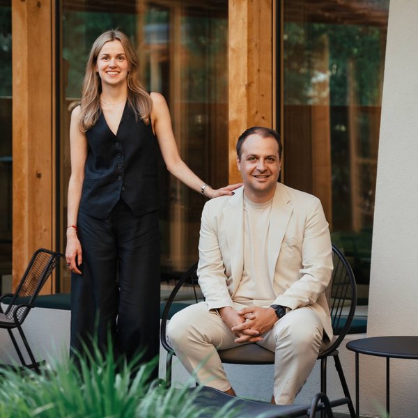 Sensoria Soulmates – Creating Memories of Bliss Frau steht, Mann sitzt auf Stuhl vor modernem Haus mit Holzrahmen