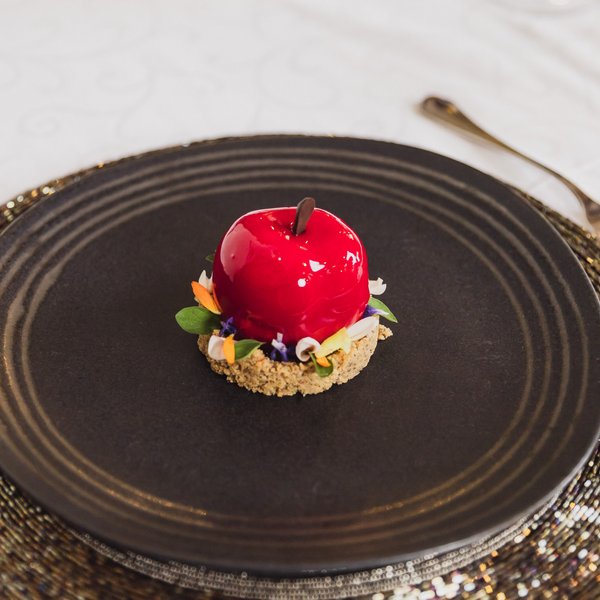 Sensoria Soulmates – Creating Memories of Bliss Shiny red apple-shaped dessert on cookie base with edible flowers on black plate